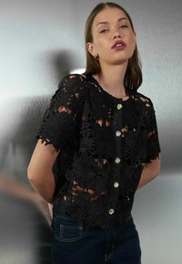 Sort broderet blonder-top med blomsterdekorationer, korte ærmer, rund halsudskæring og pyntelige knapper. Båret over mørke denimjeans.