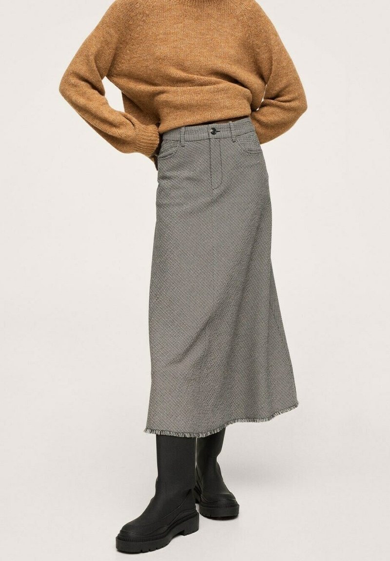 Jupe midi à motif pied-de-poule gris avec un ourlet effiloché, poches latérales et passants pour ceinture, associée à un pull en maille marron et des bottes plateformes noires.