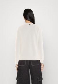 Sudadera de color crema con cuello redondeado, puños acanalados y un pequeño logotipo en la parte posterior. También se puede ver un par de pantalones cargo oscuros.