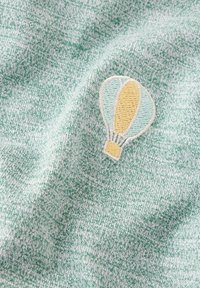 Tela verde y blanca moteada con un diseño de globo aerostático bordado que presenta colores azul, amarillo y marrón, exhibiendo una textura suave.