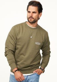 Olive green sweatshirt with ribbed texture, crew neck, long sleeves, and a small embroidered logo on the left chest. Worn with blue jeans.