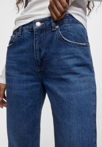 Vaqueros de denim en azul oscuro con costuras en contraste, diseño estándar de cinco bolsillos y un botón de metal en la parte superior. La textura parece ligeramente desgastada.