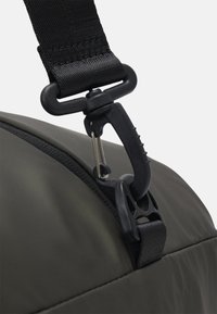 Cinta de transporte preta em nylon com um clip de plástico, presa a uma bolsa de tecido verde-escuro, apresentando uma textura suave e detalhes de costura visíveis.