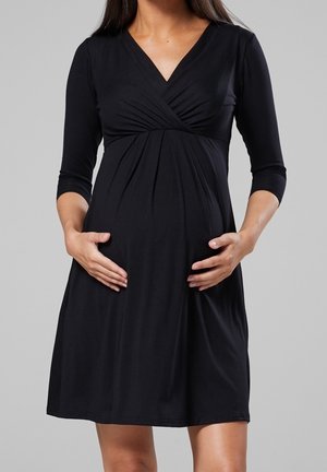 Femme enceinte portant une robe de maternité noire, mi-longue aux manches longues et col en V, tenant délicatement son ventre.