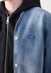 Denim jacket in light blue with silver snap buttons and a blue "HUGO" logo patch on the chest, layered over a black zip-up hoodie.