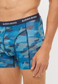 Blaue Boxershorts mit Camouflage-Muster und einem Marineblauen Bund, auf dem das Logo "BJÖRN BORG" zu sehen ist. Hergestellt aus dehnbarem Material und mit enger Passform.