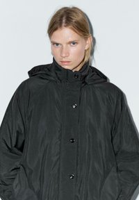 Parka nera con cappuccio e finitura testurizzata, dotata di un alto colletto, chiusura con bottoni e tasche laterali. Gli accenti includono bottoni lucidi.