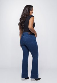 Donkerblauwe uitlopende jeans met een gladde afwerking, hoge taille en achterzakken. Model draagt een strakke zwarte top en zwarte hoge hakken.