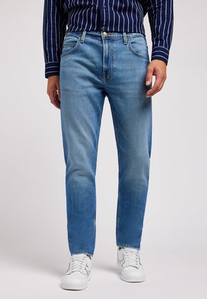 Jeans in denim azzurro chiaro con taglio dritto, sfumature sottili, design a cinque tasche e chiusura con bottone in ottone, abbinati a sneakers bianche.