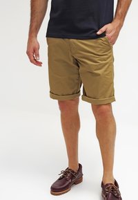 Shorts en coton beige avec des revers roulés, poches avant et coupe droite. Associés à des chaussures bateau à lacets marron foncé.
