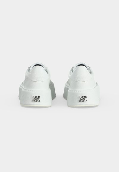 Paire de baskets basses blanches avec des semelles épaisses et un petit logo noir à l’arrière de chaque talon, sur un fond clair uni.