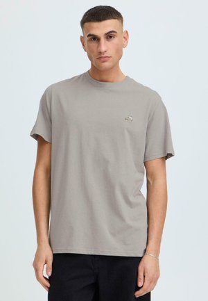 Ung mann med kort hår som har på seg en lys grå t-skjorte med et lite brodert motiv på venstre bryst, kombinert med svarte bukser.