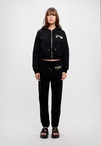 Schwarze, verkürzte Kapuzenjacke mit durchgehendem Reißverschluss und Taschen; passende schwarze Jogginghose mit grünem grafischen Detail. Getragen mit klobigen schwarzen Sandalen.