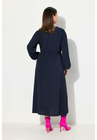 Marineblaues Langarmkleid mit gerafftem Bund, bodenlanger Schnitt, kombiniert mit lebhaft pinkfarbenen Knöchelstiefeln und glänzender Stofftextur.