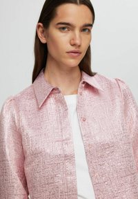 Růžová strukturovaná košile s límečkem a zapínáním na knoflíky. Materiál má lesklý, krabatý design. Nositá přes bílý top.