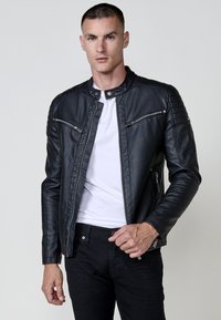 Un homme portant une veste en cuir noire sur un t-shirt blanc et un pantalon noir, se tenant contre un fond clair.