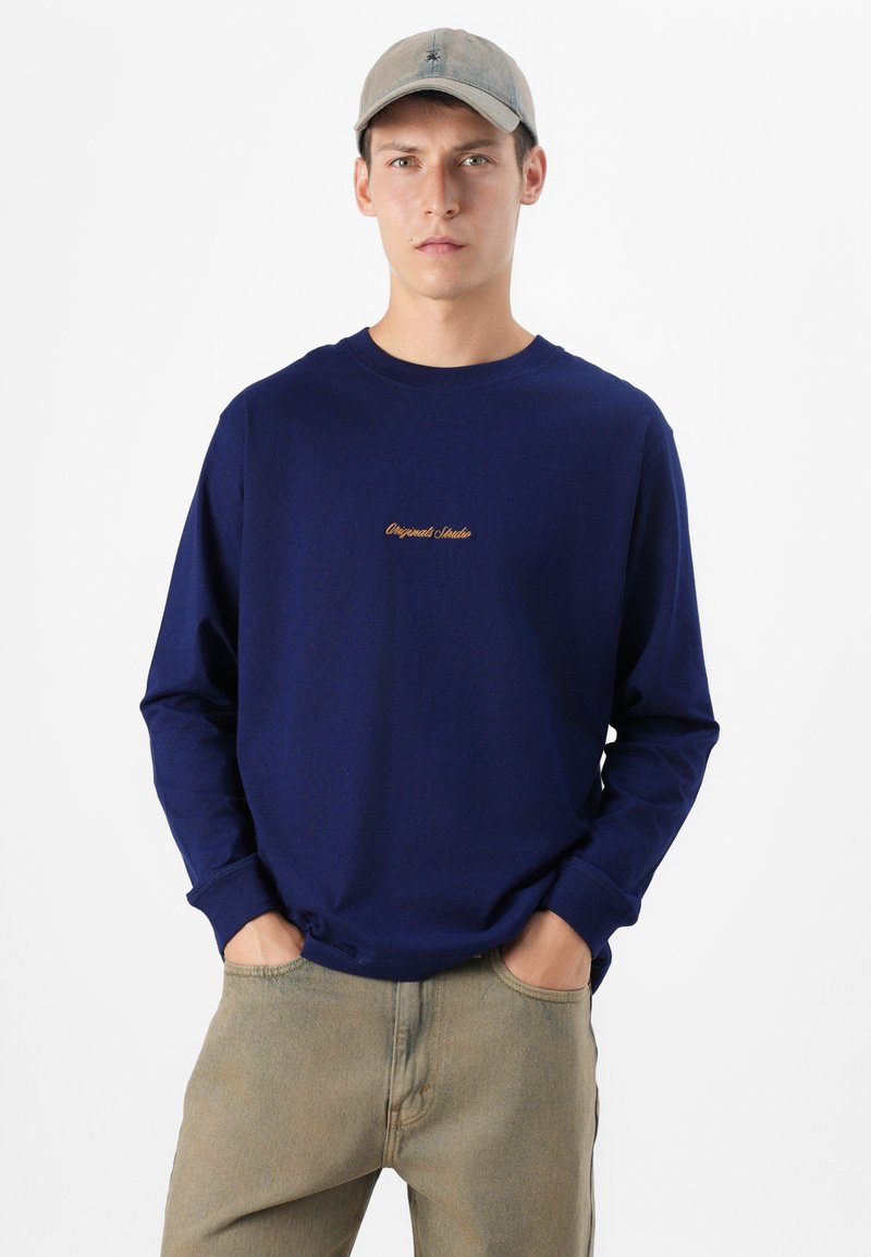 Młody mężczyzna stoi pewnie, ubrany w granatową koszulę z długim rękawem z napisem "Original Studio" i jasnymi spodniami, z rękami w kieszeniach.