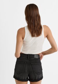 Femme aux cheveux bruns mi-longs portant un haut court sans manches blanc et un short en jean noir, vue de dos sur un fond blanc.