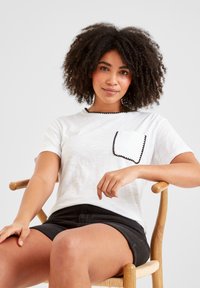 Young woman with curly hair wearing a white t-shirt with black trim and black shorts sitting on a wooden chair against white background.