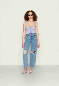 Top cropped canelado lavanda com frente amarrada, jeans claros azuis desfiados e chinelos lavanda. Modelo usa óculos de sol pretos. Fundo simples.