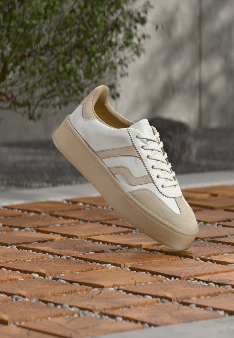 Basket avec une tige en cuir crème, des accents couleur beige foncé et une semelle en caoutchouc beige clair. Dispose de lacets plats et de détails subtils du logo sur le côté.