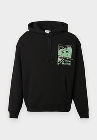 Hanorac negru cu glugă, dotat cu buzunar frontal și un grafic verde ce reprezintă un teren de tenis și copaci, inscripționat cu "Lacoste Tennis Heritage" pe piept.