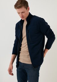 Navy blue fleece jacket with a high collar, zippered front, and side pockets. Features a chest pocket with a zip and soft texture.