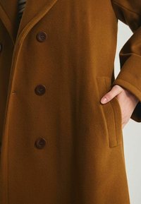 Manteau en laine brun avec un design croisé, de grands boutons ronds et des poches latérales avec une finition douce et texturée.