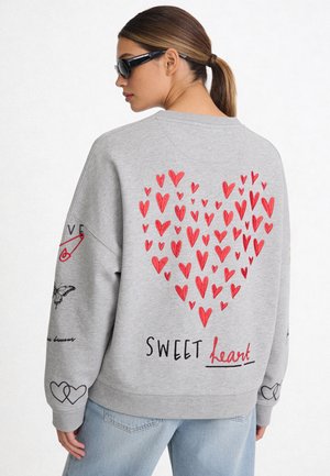 Femme portant un sweatshirt gris avec un groupe de cœurs rouges et le texte "sweet heart" dans le dos, associé à un jean bleu clair et des lunettes de soleil noires.