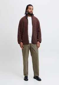 Bruine fleece ritssluiting jack met hoge kraag, gecombineerd met een wit shirt, olijfgroene broek en zwarte schoenen. Twee zijzakken zichtbaar.