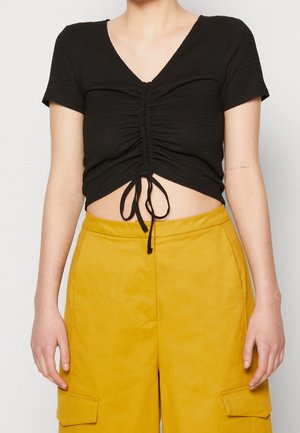 Persona que lleva un crop top negro fruncido con mangas cortas y pantalones mostaza de cintura alta con bolsillos.