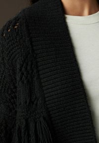Cardigan negru tricotat cu o textură ribbed, având designuri de tricot deschis pe umeri și detalii cu ciucuri la tiv. Poartă-l peste o bluză deschisă.