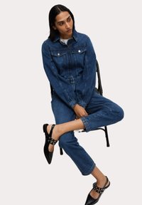 Denimová kombinéza v tmavě modré barvě s dlouhými rukávy, předními kapsami a volným střihem; doplněna černými slingback botami s ozdobnými sponami.