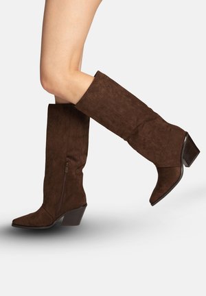 Kvinde iført mørkebrune ruskindsstøvler til knæet med sideslynge og blokhæl, et ben løftet mod hvid baggrund.