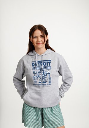 Młoda kobieta o brązowych włosach ma na sobie szarą bluzę z kapturem z grafiką Detroit oraz zielone, paskowane szorty, stoi z rękami za plecami.
