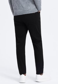 Pantalon noir tapered avec une finition en tissu lisse, doté d'une poche arrière et de poignets élastiques, associé à des baskets blanches sur un fond uni.