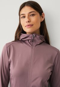 Giacca viola chiaro con colletto alto e zip frontale. Tessuto liscio e leggero con dettagli delle cuciture e una vestibilità aderente sulle spalle.
