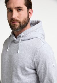 Hellgrauer Kapuzenpullover aus weichem Material. Mit Kordelzug, einer vorderen Kängurutasche und einem Logo-Patch am Ärmel.