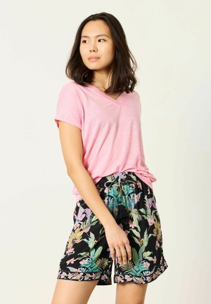 Młoda kobieta z ciemnymi włosami do ramion, ubrana w różowy top z krótkim rękawem i dekoltem w serek oraz czarne szorty w kwiatowy wzór, stojąca i patrząca w lewo.