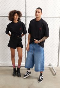 Sorte oversized t-skjorter med røde leppebroderier, sort miniskjørt med belte, blå vide bukser og sorte støvler. Chain-link bakgrunn.