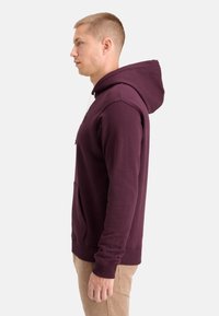 Burgunderfarbene Hoodie aus Baumwollmix, ausgestattet mit einer Fronttasche, verstellbarem Kordelzug an der Kapuze sowie Rippenbündchen und -saum. Seitenansicht.