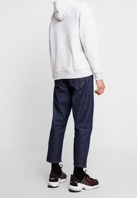 Person som bär en ljusgrå hoodie, mörkblå jeans som slutar ovanför anklarna, svarta strumpor och svarta Adidas-sneakers med röda inslag, stående mot en vit bakgrund.