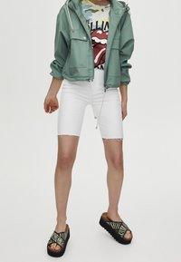 Chaqueta impermeable verde, camiseta gráfica con el logo de los Rolling Stones, pantalones cortos de mezclilla blancos, sandalias negras con diseño a rayas y suelas gruesas.