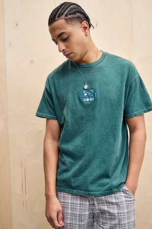 Jeune homme avec des cheveux tressés portant un T-shirt vert avec un écusson de poisson et un short à carreaux, se tenant contre un fond en bois clair.