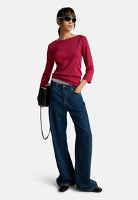 Frau trägt dunkle Sonnenbrille, ein rotes Langarmoberteil, weite blaue Jeans, schwarze Schuhe und hält eine schwarze Handtasche.