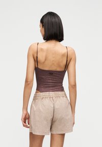 Γυναίκα με κοντά μαύρα μαλλιά, που φοράει καφέ αμάνικο τοπ και μπεζ σορτς με ελαστική μέση, στέκεται γυρισμένη με την πλάτη προς το απλό φόντο.