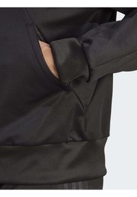 Suéter negro con un acabado texturado, que presenta un bolsillo lateral y puños acanalados. El diseño enfatiza un ajuste elegante y atlético.