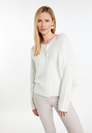 Cardigan alb cu textura ribbed, cu un fit lejer, având guler rotund, închidere cu nasturi și mâneci lungi, asortat cu pantaloni bej deschis.