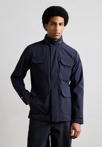 Άνδρας με αφροκεντρικό χτένισμα που φοράει ένα σκούρο μπλε μπουφάν με τέσσερις μπροστινές τσέπες, στέκεται μπροστά σε απλό ανοιχτό γκρι φόντο.