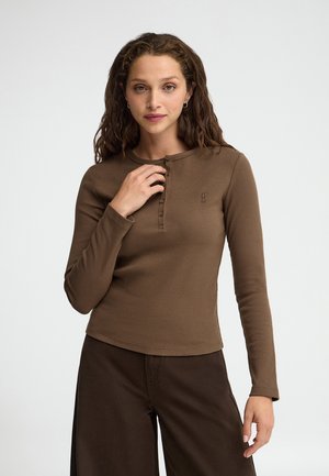 Mujer con pelo rizado que lleva una camiseta henley de manga larga marrón y pantalones marrón oscuro, de pie frente a un fondo claro y liso.